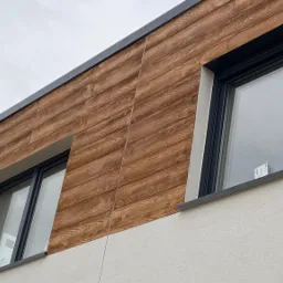 Fragment elewacji domu z drewnopodobnymi panelami i czarnymi ramami okiennymi, widoczne dwa okna i fragment białego tynku na dole.
