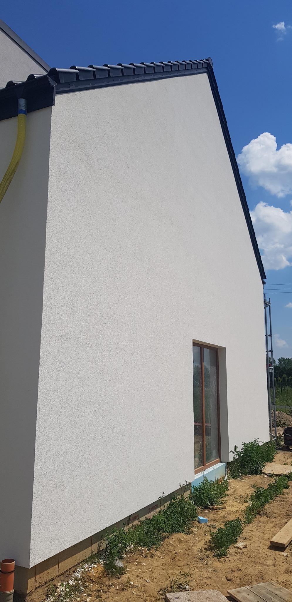 Fragment ściany budynku z nową białą elewacją o strukturze baranka, widoczne okno z brązową ramą, czarne orynnowanie i fragment dachu z dachówką, błękitne niebo w tle.