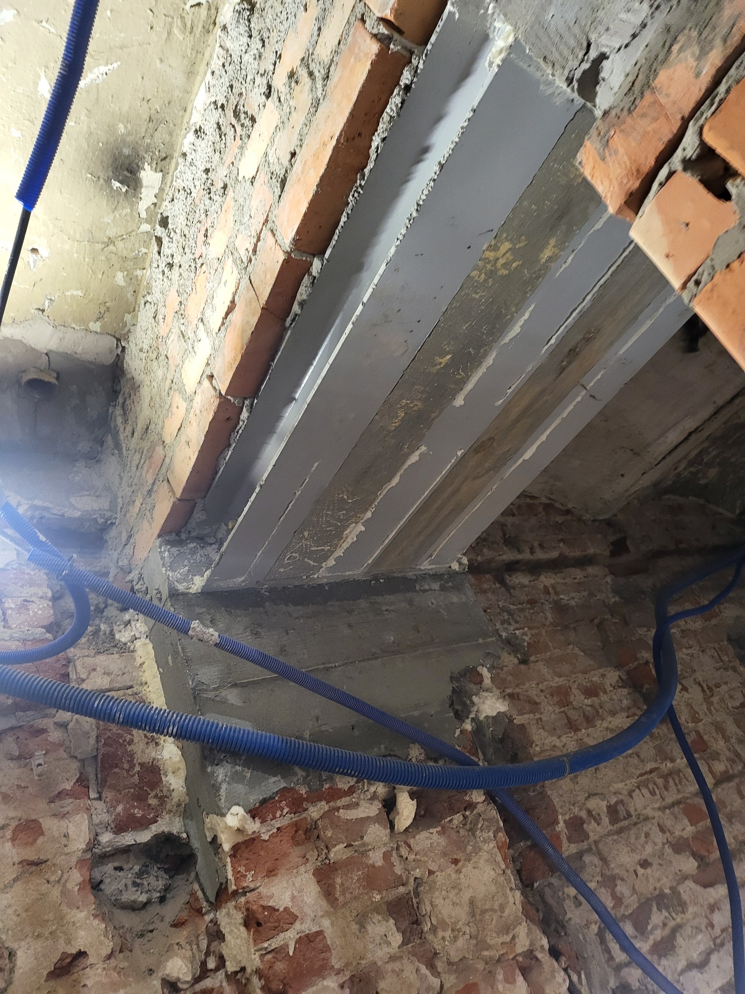 Ceglany mur z szarymi belkami wzmacniającymi i niebieskimi rurami instalacyjnymi. Widoczne ślady remontu i surowy stan wykończenia wnętrza.