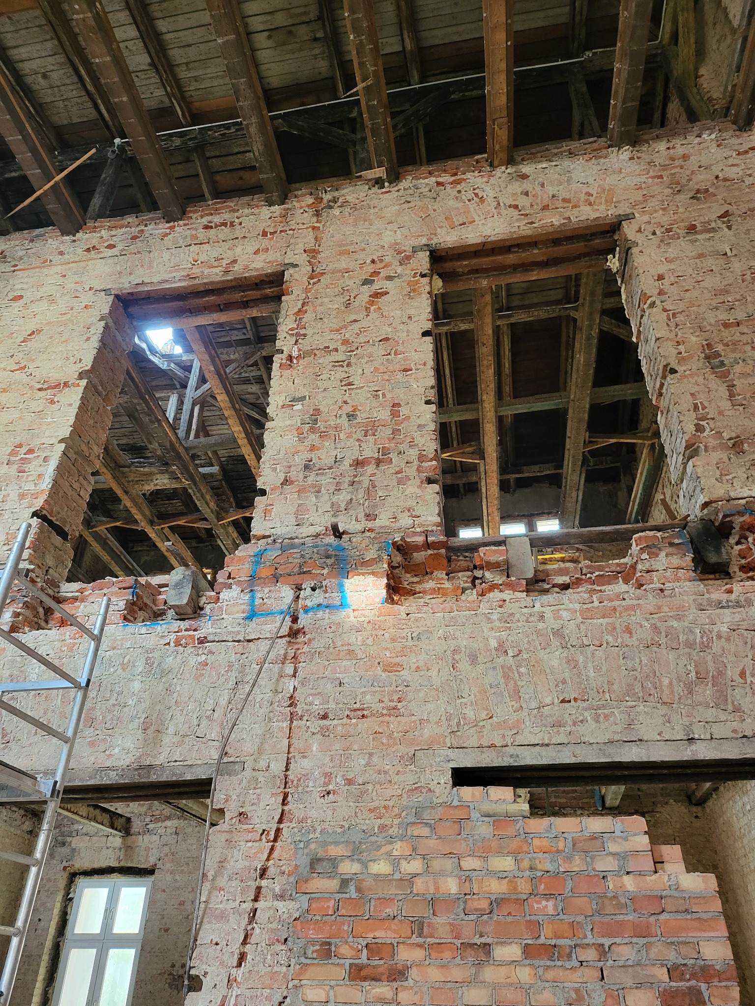Ceglana ściana z otworami okiennymi w trakcie remontu. Widoczne drewniane elementy konstrukcyjne i fragment rusztowania. Częściowo usunięty tynk odsłania strukturę cegieł.