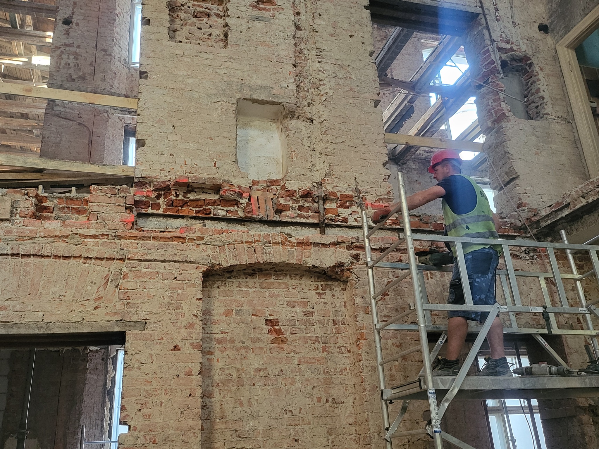 Murarz na rusztowaniu, w kasku, pracuje przy renowacji ceglanej ściany wewnątrz budynku. Widoczne fragmenty konstrukcji dachu i okna. Prace renowacyjne w toku.