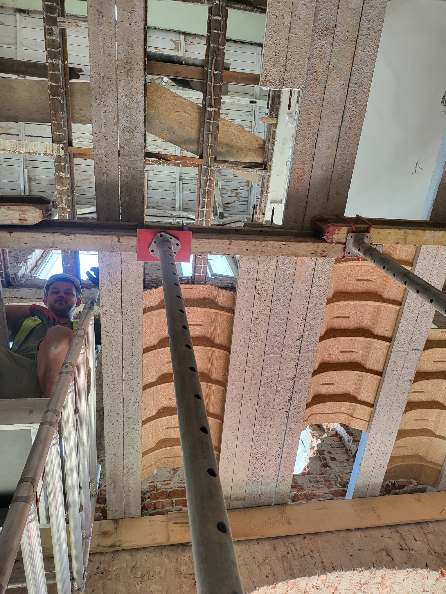 Prace remontowe na poddaszu: pracownik na drabinie, widoczne elementy konstrukcyjne stropu i ściany z cegły, podpory stalowe. Ujęcie z dołu, remont domu.