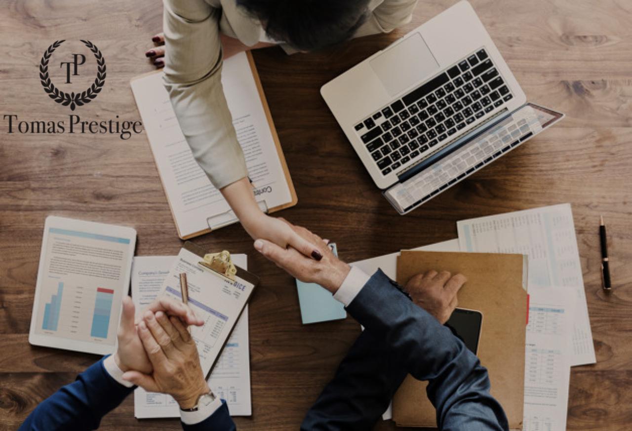 Widok z góry na drewniany stół z laptopem, tabletem z wykresami, dokumentami, długopisem i dłońmi dwóch osób ściskających się na znak zawarcia umowy. Na stole widoczne logo firmy Tomas Prestige.