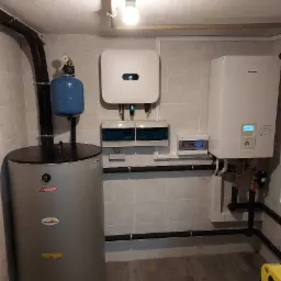 Kotłownia z szarym bojlerem Termica inoxsolar, niebieskim zbiornikiem wyrównawczym, białym inwerterem Huawei i kotłem gazowym Panasonic na tle jasnoszarych płytek i parkietu.