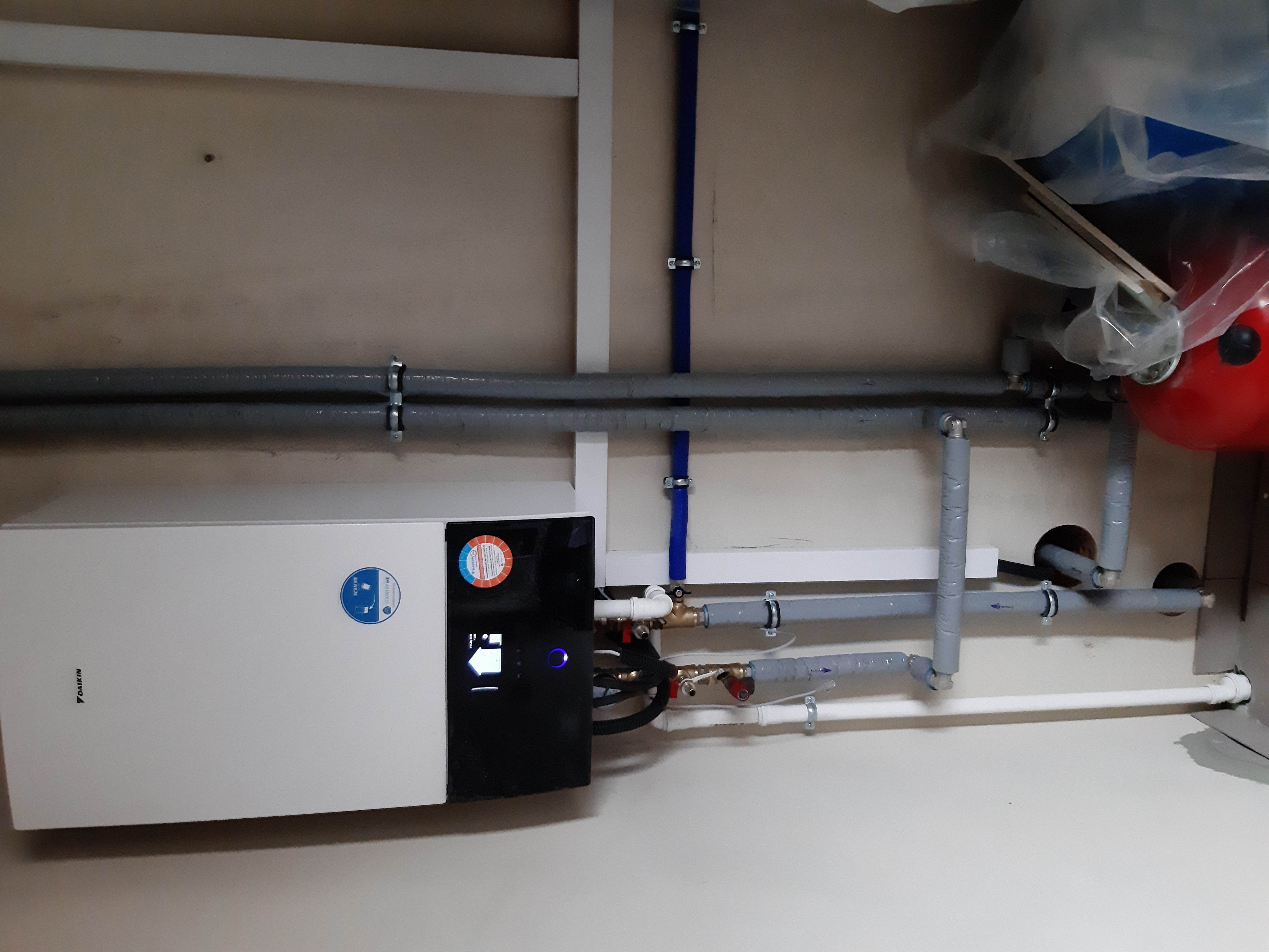 Biała pompa ciepła Daikin z podłączonymi rurami instalacji hydraulicznej, widoczny zbiornik wyrównawczy i fragment instalacji w pomieszczeniu technicznym.