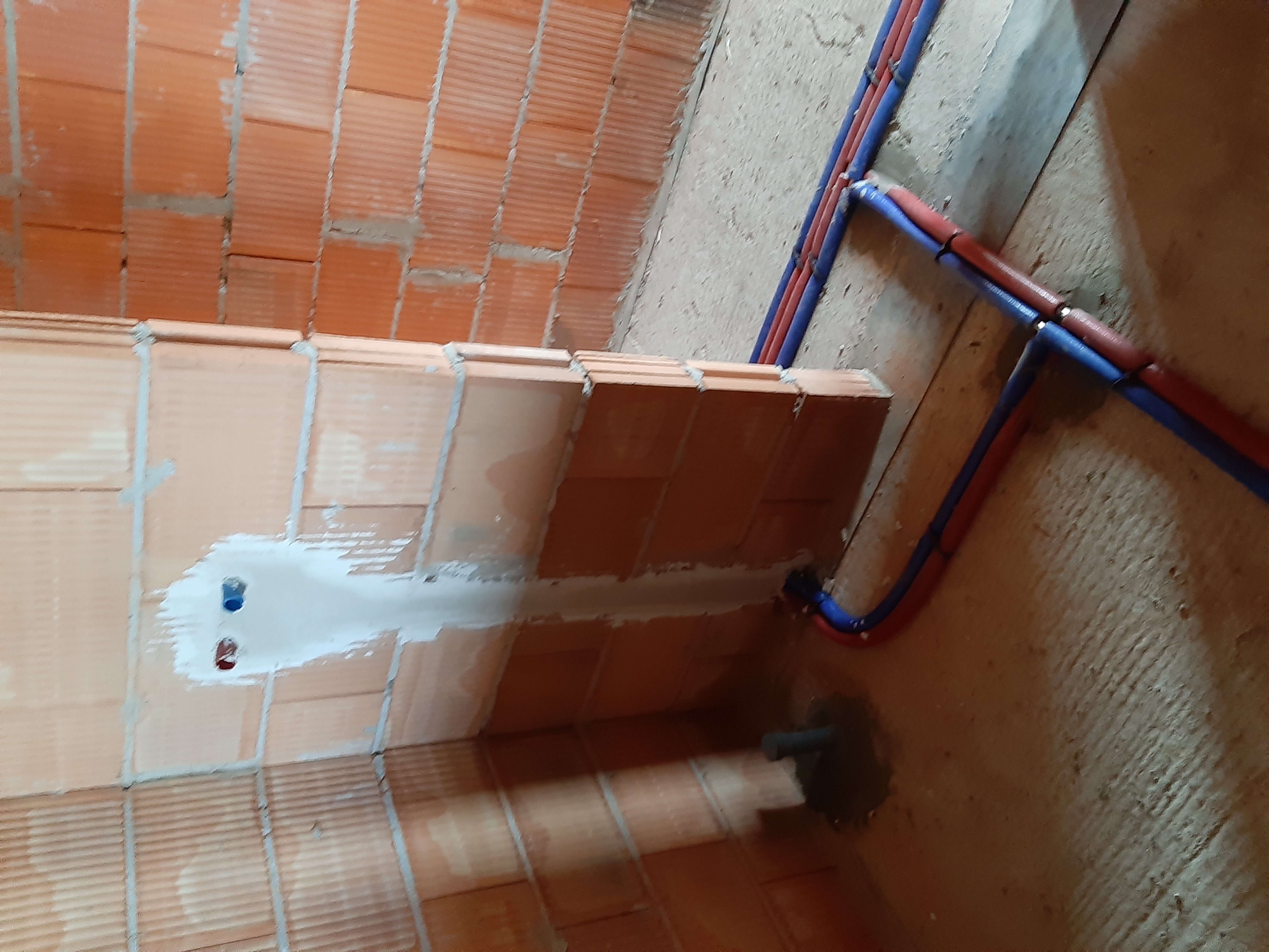 Ściana z pustaków z wyciętymi otworami na instalację wodną i częściowo zamurowanymi rurami, widoczne fragmenty instalacji z rur czerwonych i niebieskich.