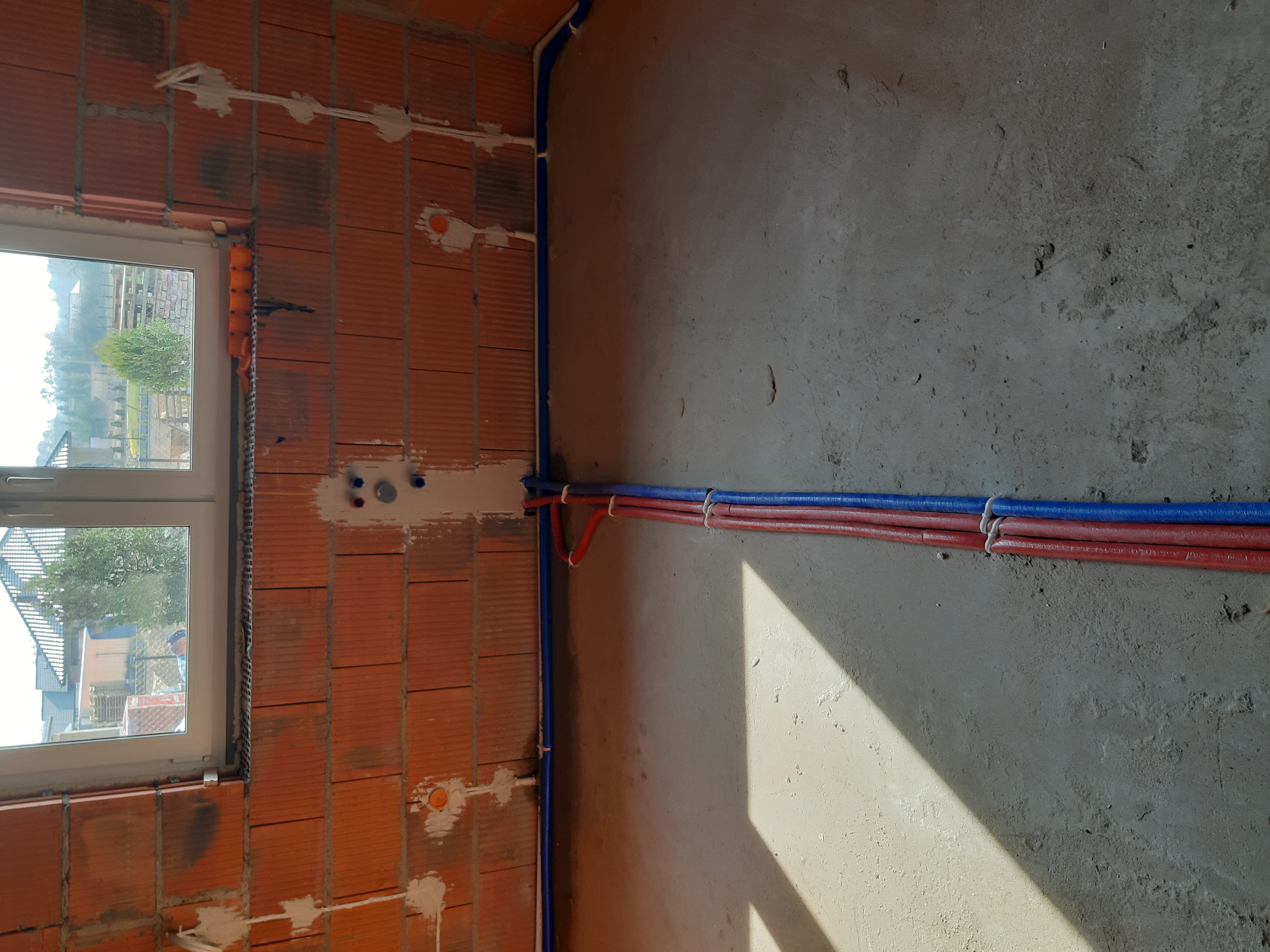 Instalacja wodna i kanalizacyjna w surowym budynku z widocznym oknem i ceglaną ścianą oraz wiązką rur czerwonych i niebieskich mocowanych do ściany betonowej.
