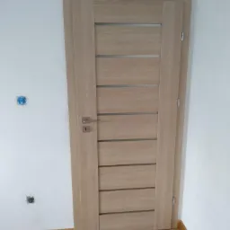 Jasne, drewniane drzwi wewnętrzne z poziomymi przeszkleniami, zamontowane w białej ścianie, widoczne gniazdka elektryczne na ścianie i drewniana podłoga.