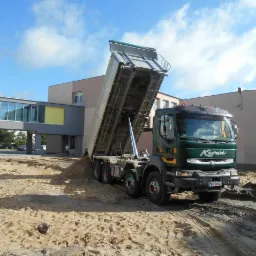 Ciężarówka Renault Kerax z podniesioną skrzynią wyładowuje materiał na placu budowy, widoczne ślady opon na piaszczystym podłożu, w tle budynek z łącznikiem.