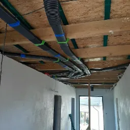 Szare przewody wentylacyjne zamontowane pod drewnianym stropem, przymocowane zielonymi uchwytami, widok z korytarza w trakcie wykańczania.