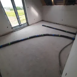 Montaż rur rekuperacji na poddaszu w surowym stanie, widoczne rury z zielonymi i niebieskimi oznaczeniami, okno z widokiem na zielony krajobraz.