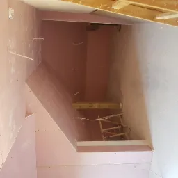 Wnętrze poddasza w trakcie prac wykończeniowych: różowe płyty na ścianach i skosach, widoczne kable, fragment rusztowania i surowa, biała ściana.