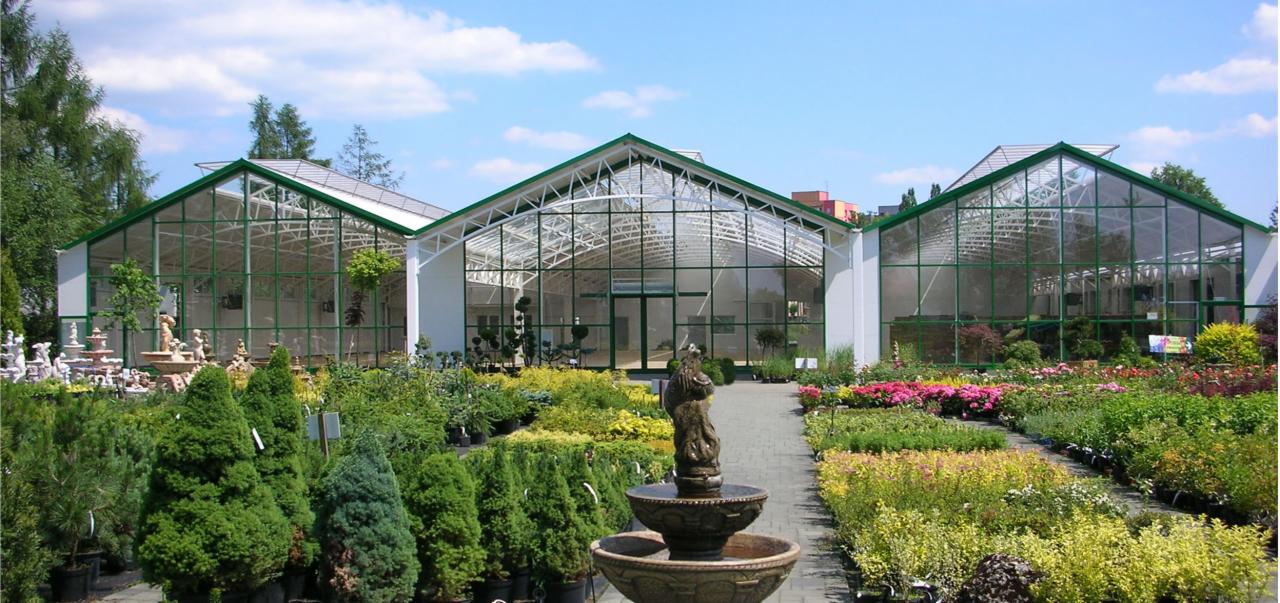 Kompleks szklarni z zielonymi ramami i przeszklonymi ścianami, otoczony ogrodem z fontanną i różnorodną roślinnością ozdobną, widok z zewnątrz w słoneczny dzień.