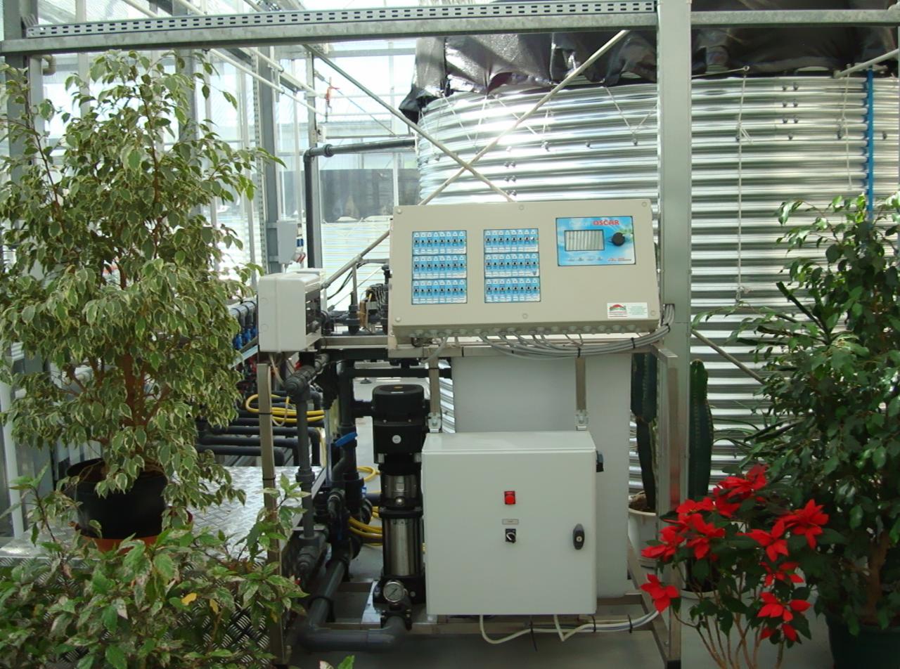 System nawadniania w szklarni z metalowym zbiornikiem na wodę, panelem sterowania i pompą, otoczony roślinami doniczkowymi.