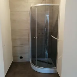 Wykończona łazienka z kabiną prysznicową p&oacute;łokrągłą, szare płytki na ścianach i ciemnobrązowa podłoga, widoczne niedokończone podłączenie elektryczne do oświetlenia sufitowego.