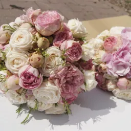 Dwa bukiety ślubne z róż w odcieniach bieli i pudrowego różu, z dodatkiem zielonych gałązek, ułożone na białym tle.