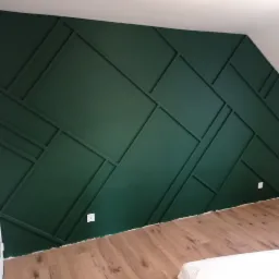 Ściana w pokoju pomalowana na ciemnozielono z dekoracyjnymi listwami tworzącymi geometryczny wzór, widoczne gniazdka elektryczne i fragment podłogi z paneli.