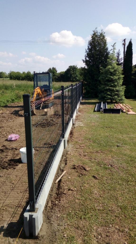 Montaż ogrodzenia panelowego na betonowej podmurówce, widoczny minikoparka w tle i przygotowane elementy ogrodzenia na trawie.