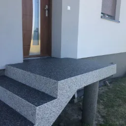 Nowoczesne schody wejściowe z szarego kamienia kompozytowego prowadzące do drewnianych drzwi wejściowych, wsparte na okrągłym filarze, z widoczną strukturą kamienia.