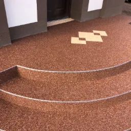 Brązowa, dwustopniowa kaskada schodów wejściowych z żywicy z metalowymi listwami krawędziowymi i dekoracyjnym wzorem z jasnych kwadratów na podwyższeniu.