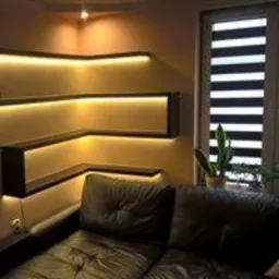 Nowoczesne półki narożne z podświetleniem LED w kolorze ciepłej bieli, umieszczone nad skórzaną sofą w salonie. Widoczna roleta rzymska w oknie i roślina doniczkowa.