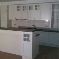 Biała zabudowa kuchenna z drewnianym blatem i charakterystycznymi przeszklonymi frontami szafek górnych w kratkę. Widoczna wyspa kuchenna z frontem z motywem kratki. Jasne płytki na ścianie. Ujęcie...