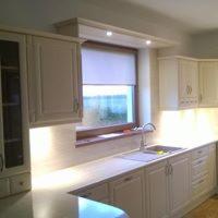 Jasna, klasyczna zabudowa kuchenna z frezowanymi frontami, oświetleniem podszafkowym LED i blatem z jasnego drewna, widoczna pod oknem roleta zwijana.