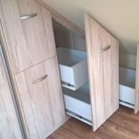 Zabudowa pod skosem z jasnego drewna, wysuwane szuflady z białymi ściankami, srebrne uchwyty, widoczna podłoga z paneli.