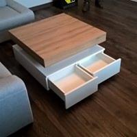 Kwadratowy stolik kawowy z jasnego drewna, osadzony na białej podstawie z dwoma wysuniętymi szufladami. Widoczna część sofy w tle.