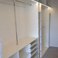 Wnętrze garderoby z białymi półkami, szufladami z metalowymi frontami i drążkiem na ubrania, oświetlenie LED w pionowych panelach.