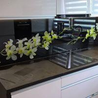 Białe fronty szafek kuchennych z blatem w odcieniach szarości, ozdobione kompozycją z białych orchidei i czarnym, prostokątnym wazonem. W tle widoczny piekarnik w zabudowie i rolety okienne.