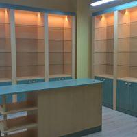 Jasne, drewniane regały z niebieskimi akcentami, oświetlone od góry, z przeszklonymi półkami i szafkami w dolnej części, widoczne za ladą z półkami, tworzące zabudowę pomieszczenia.