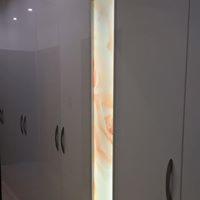 Biała zabudowa z połyskiem, oświetlona pionowym panelem z efektem marmuru, widoczne uchwyty szafek.