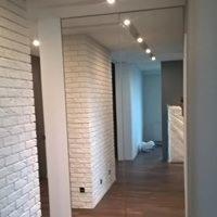 Wnętrze korytarza z dużą, lustrzaną szafą wnękową, optycznie powiększającą przestrzeń. Ściana wykończona białą cegłą dekoracyjną, podłoga z ciemnego drewna. Widoczne oświetlenie punktowe na suficie.