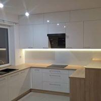 Biała zabudowa kuchenna w kształcie litery U z jasnymi blatami, oświetleniem LED pod szafkami wiszącymi i czarnym okapem.