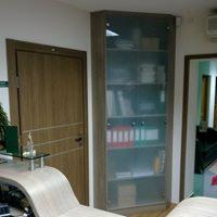 Wysoka, narożna szafa z przeszklonymi drzwiami, wbudowana w rogu pokoju, widoczna obok drewnianych drzwi i fragmentu blatu biurka. Wewnątrz szafy widoczne segregatory i inne dokumenty.