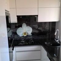 Białe szafki kuchenne z czarnym blatem i panelem szklanym z motywem białej orchidei nad płytą grzewczą i zlewem.