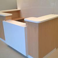 Recepcja z jasnego drewna i białych paneli, zaokrąglone krawędzie, minimalistyczny design, widoczne delikatne refleksy światła na powierzchni.