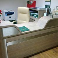 Biurko z jasnego drewna z zaokrąglonymi elementami, zielony notatnik na blacie, krzesło z jasną tapicerką w tle, drukarka i kalendarz na ścianie.