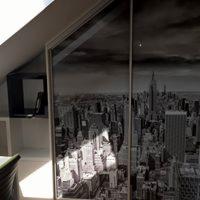 Biała szafa przesuwna na poddaszu ze szklanym frontem z czarno-białym motywem panoramy Nowego Jorku, widoczna półka i fragment biurka po lewej stronie.