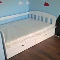 Białe łóżko z szufladami, materac owinięty folią, ustawione w rogu pokoju z niebieskimi ścianami i dekoracjami chmur.