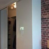 Biała, minimalistyczna zabudowa z przesuwnymi drzwiami, wnęka z półkami i wieszakami widoczna po otwarciu, ceglana ściana w tle.