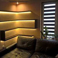 Nowoczesne półki narożne z podświetleniem LED w kolorze ciepłej bieli, umieszczone nad skórzaną sofą w salonie. Widoczna roleta rzymska w oknie i roślina doniczkowa.