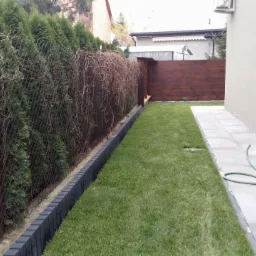 Uporządkowany ogród z równo przystrzyżonym trawnikiem, obrzeżem z ciemnych kostek i żywopłotem z tuj wzdłuż ogrodzenia z siatki, częściowo oplecionego suchymi pnączami. W tle widoczny fragment...