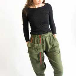 Modelka prezentuje zielone spodnie dresowe z naszytą kieszenią i czerwonymi paskami, czarna bluzka z długim rękawem, białe trampki, okulary na głowie, białe tło.