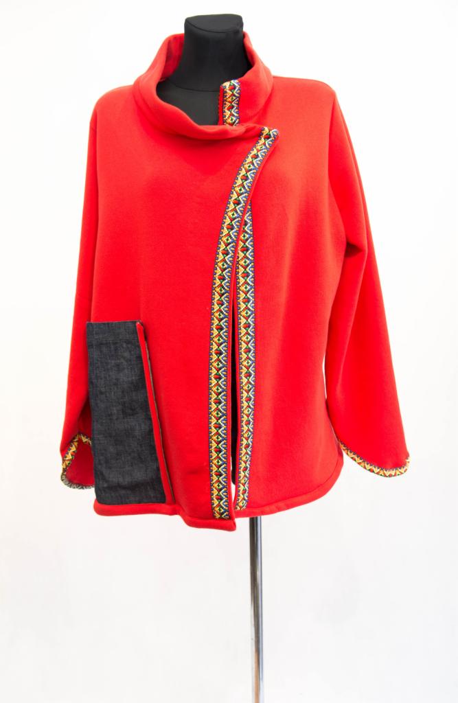 Czerwona bluza damska z asymetrycznym zapięciem, ozdobiona kolorową, etniczną taśmą i ciemną, dżinsową kieszenią, prezentowana na manekinie na białym tle.