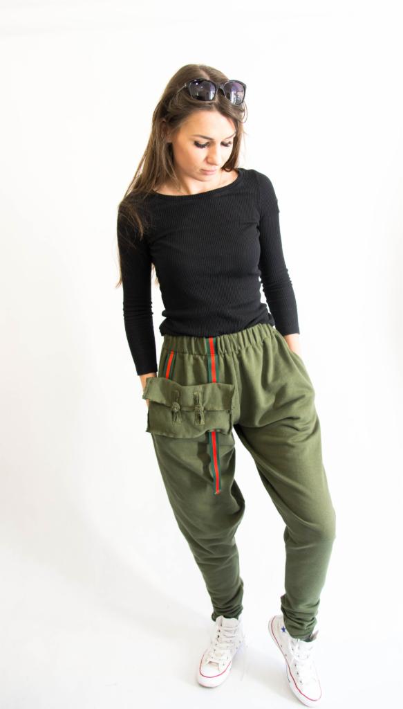 Modelka prezentuje zielone spodnie dresowe z naszytą kieszenią i czerwonymi paskami, czarna bluzka z długim rękawem, białe trampki, okulary na głowie, białe tło.