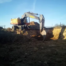 Żółto-szara koparka kołowa Ackermann podczas wykopu w ziemi o gliniastej strukturze, na tle błękitnego nieba i wczesnowiosennej zieleni.