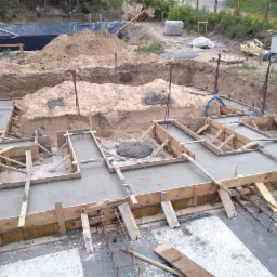 Wylane fundamenty z drewnianym szalunkiem i wystającymi prętami zbrojeniowymi, widoczne wykopy i hałda piasku w tle.