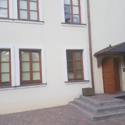 Fragment elewacji budynku z kremową fasadą, brązowymi oknami z białymi ramami, wejściem z zadaszeniem i schodami wyłożonymi kostką brukową